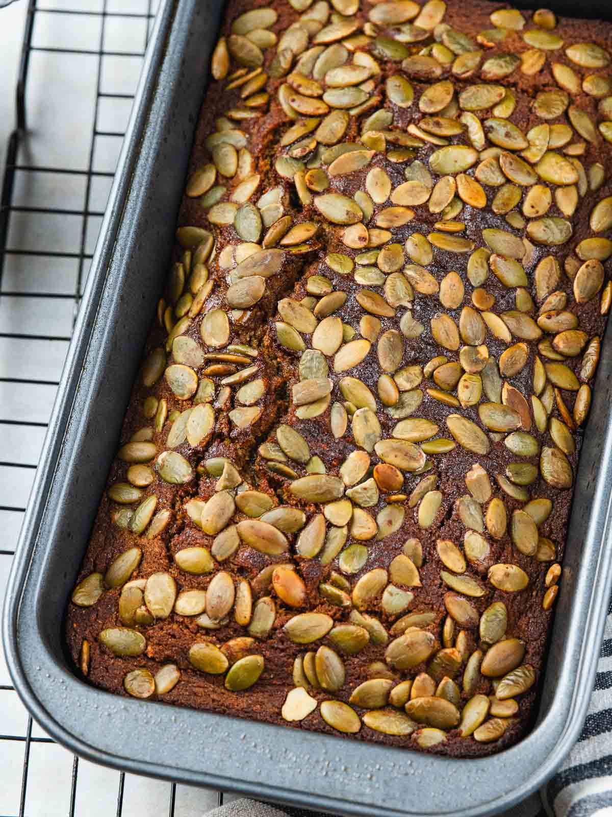 Loaf of quick bread topped with pumpkin seeds in a metal baking pan on a wire rack.