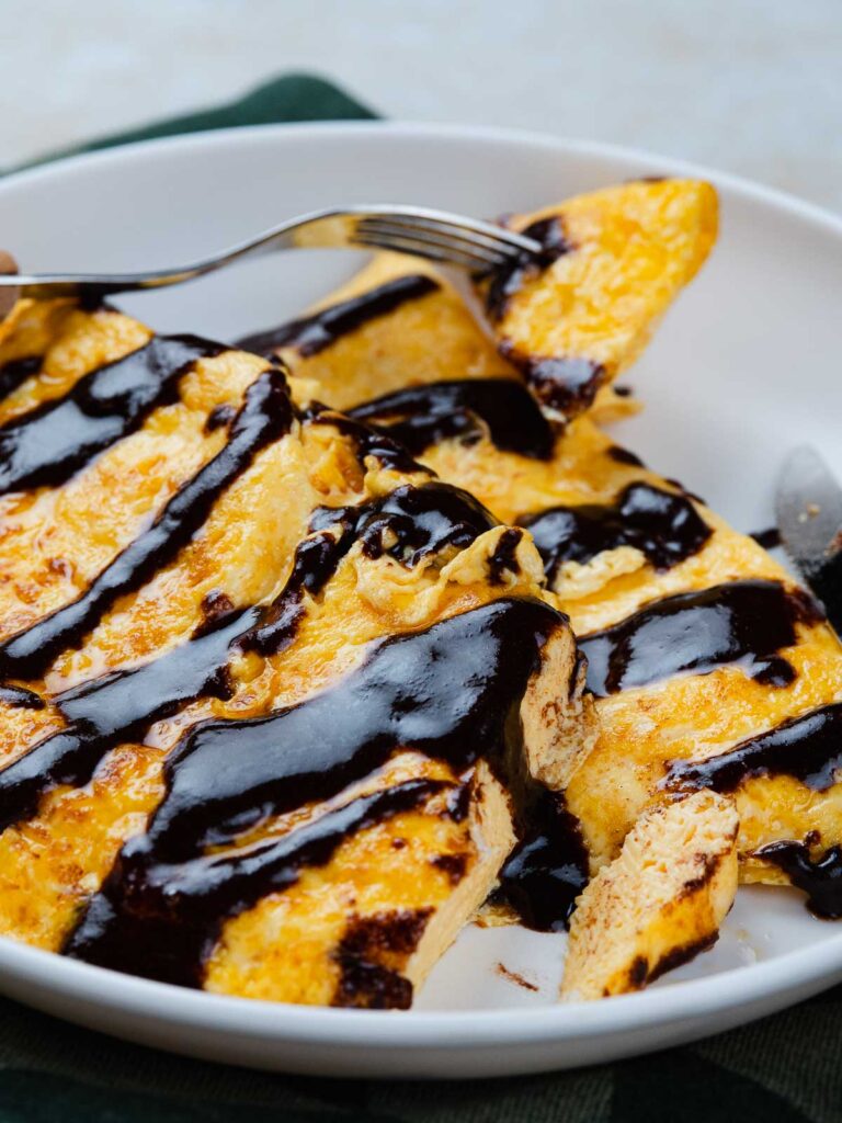 A plate of cooked egg topped with dark chocolate sauce, being eaten with a fork.