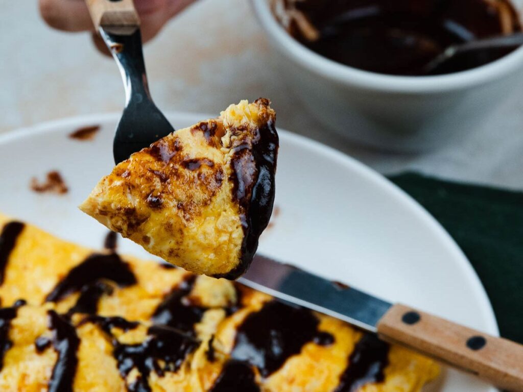 A fork holding a piece of omelet drizzled with dark sauce, positioned over a plate with more of the omelet.