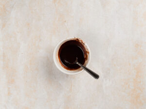 A small white bowl containing dark liquid chocolate with a metal spoon inside.