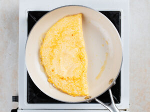 A folded omelet cooking in a light-colored frying pan on a stovetop.
