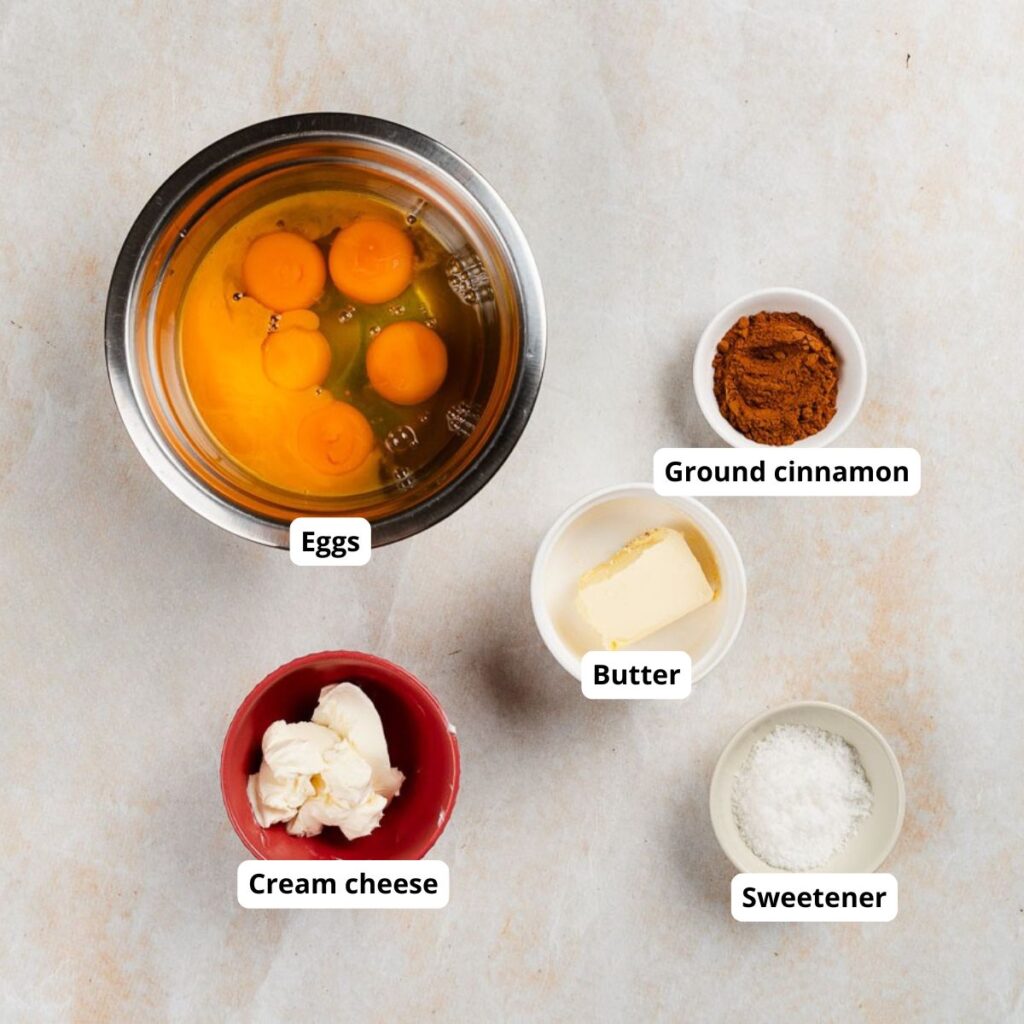 Ingredients for a recipe in bowls: eggs in a metal bowl, and cream cheese, butter, ground cinnamon, and sweetener in small individual bowls.