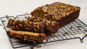 A loaf of pumpkin seed bread on a wire cooling rack, with two slices cut from the end.