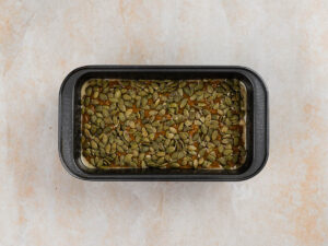 A rectangular loaf pan filled with dough topped with a layer of green pumpkin seeds.
