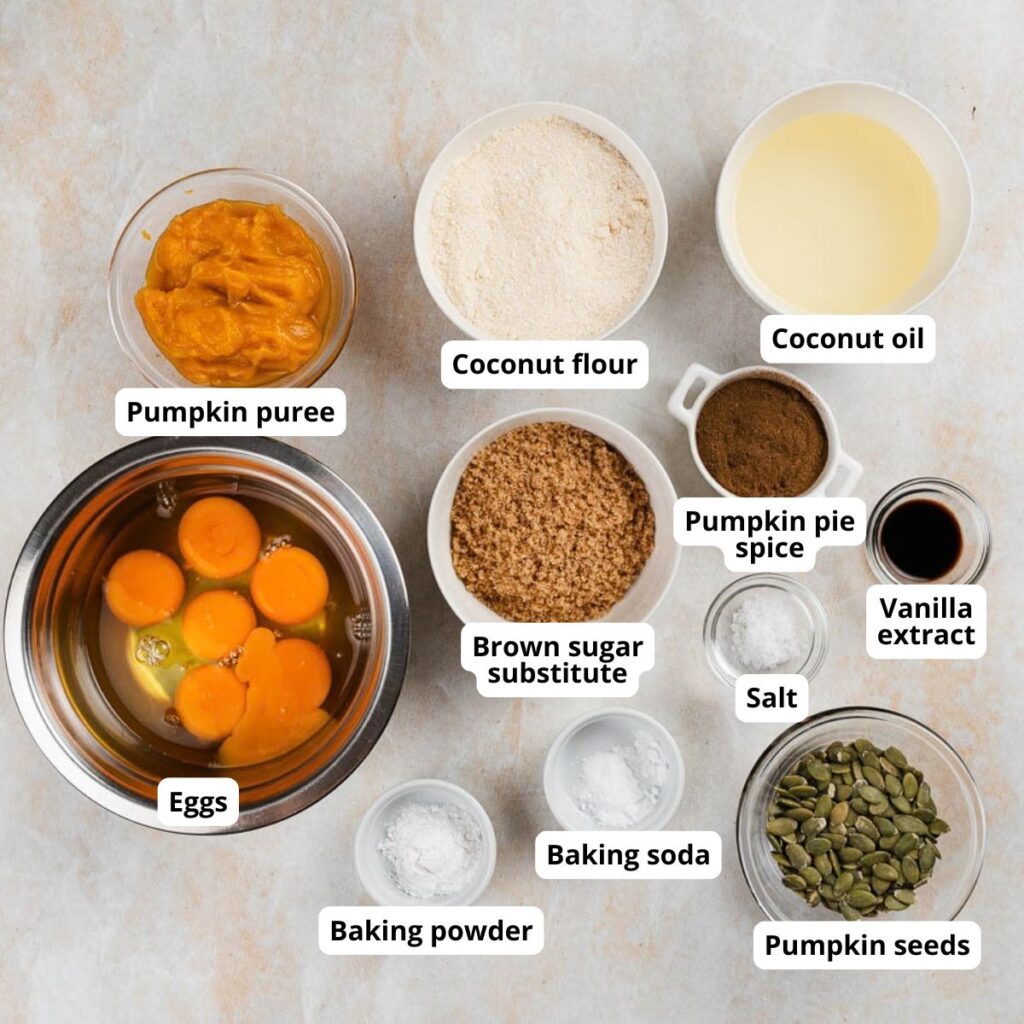 Top-down view of ingredients for a pumpkin recipe, labeled: pumpkin puree, coconut flour, coconut oil, eggs, brown sugar substitute, pumpkin pie spice, vanilla extract, salt, baking soda, baking powder, and pumpkin seeds.