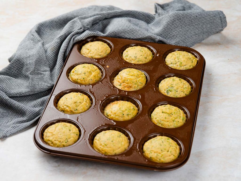 A brown muffin tin filled with eleven baked savory muffins next to a gray cloth.