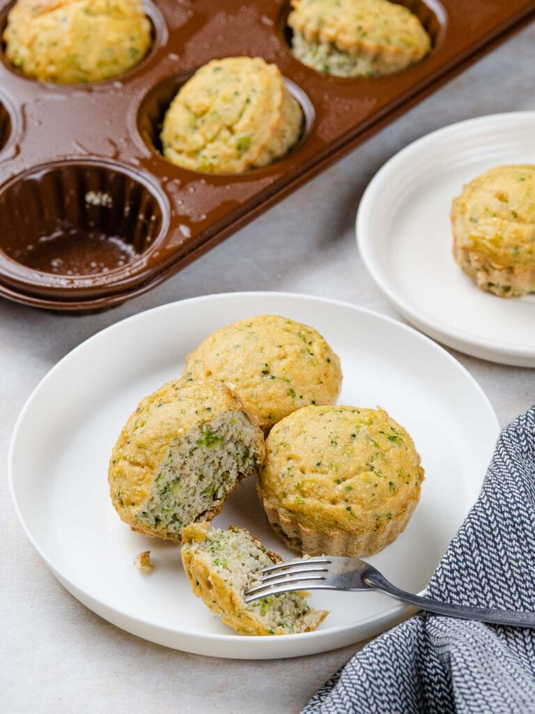 Savory broccoli muffins in a muffin tin and on white plates, with one broken open by a fork.