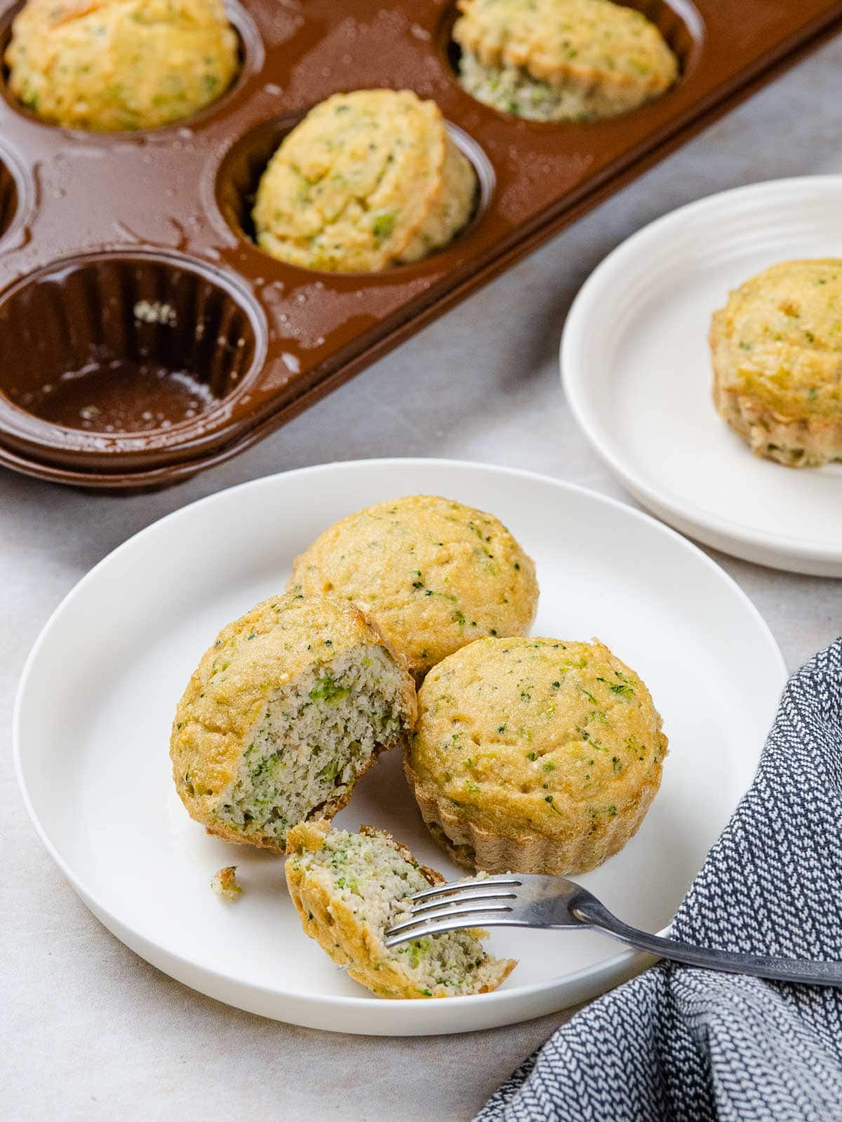 Savory broccoli muffins in a muffin tin and on white plates, with one broken open by a fork.