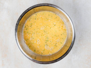 A metal mixing bowl filled with whisked eggs and finely chopped green vegetables.
