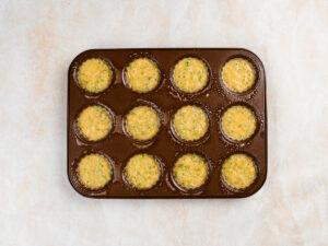 A muffin tin filled with twelve scoops of raw broccoli and egg batter.